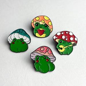 Può includere: Quattro spille smaltate raffiguranti rane verdi sotto cappelli di funghi colorati. Una rana tiene un cuore, un'altra suona l'ukulele e le altre hanno diversi disegni di funghi. Le spille hanno uno sfondo bianco.