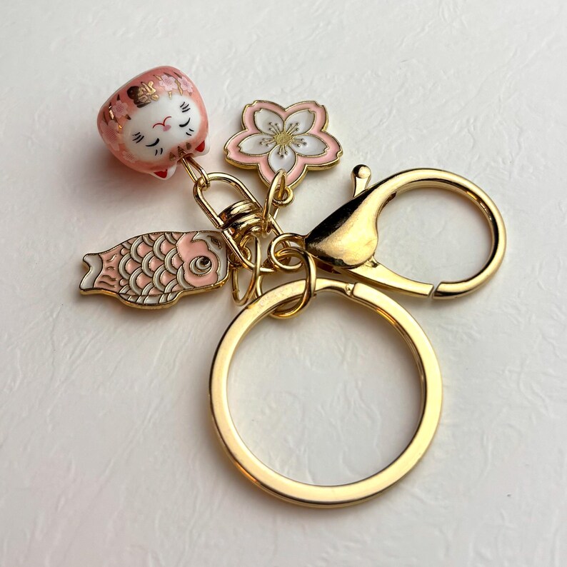 May include: A gold-colored keychain featuring Japanese-inspired charms. The charms include a pink and white lucky cat, a cherry blossom flower, and a koi fish. The keychain has a large ring and a clasp for attaching to keys or a bag.