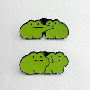 Puede incluir: Dos pines de esmalte con ranas de dibujos animados verdes. Un pin muestra dos ranas besándose, el otro muestra dos ranas sonriendo. Los pines tienen contornos negros y están sobre un fondo blanco.