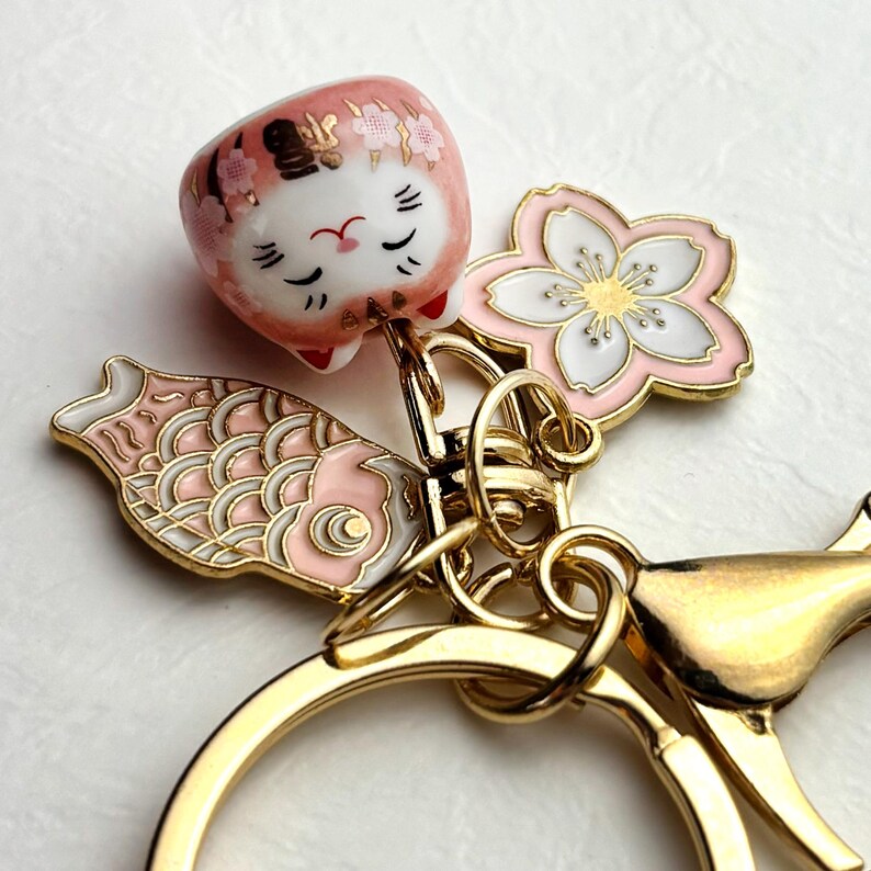 May include: A gold-toned keychain featuring a pink and white lucky cat charm, a pink and white cherry blossom, and a pink and white koi fish charm. The lucky cat has black facial details and the word "福" on its head.