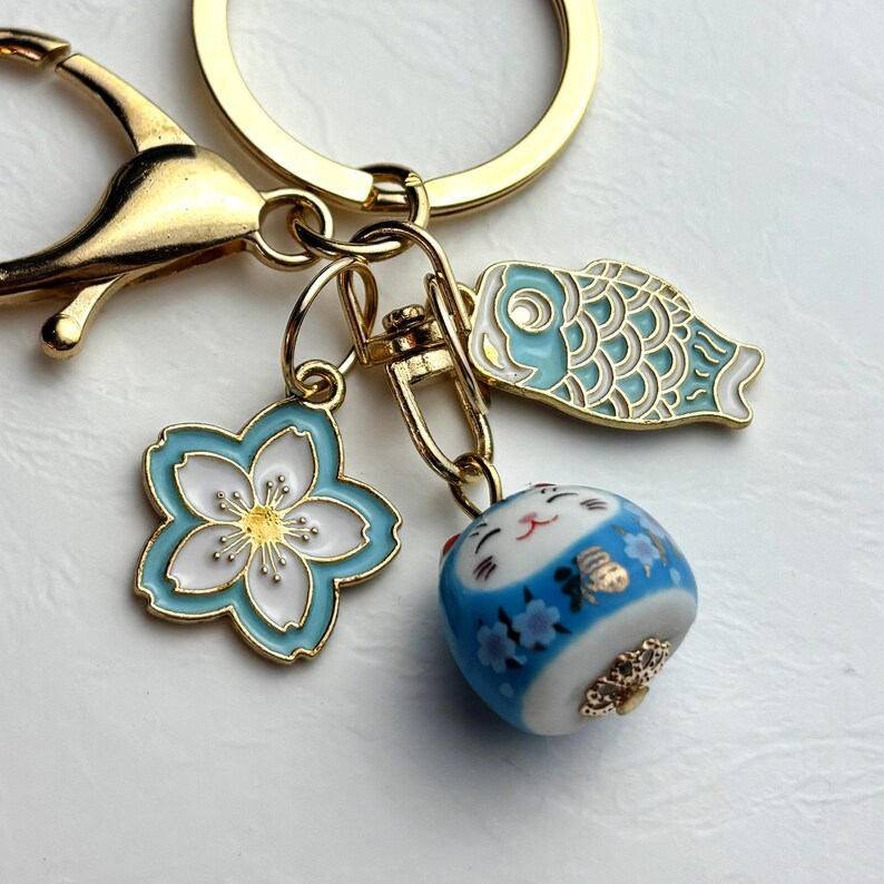 May include: Gold-tone keychain with three charms: a blue and white cherry blossom, a blue and white fish, and a blue and white lucky cat figurine. The keychain has a lobster clasp and a split ring.