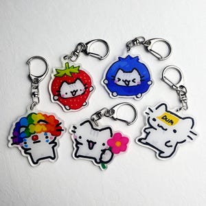 May include: A collection of acrylic keychains featuring cartoon cat characters. Each keychain has a silver clasp and a unique design, including a strawberry, blueberry, flower, and a rainbow-colored clown. One keychain has the word "DUM" on it.
