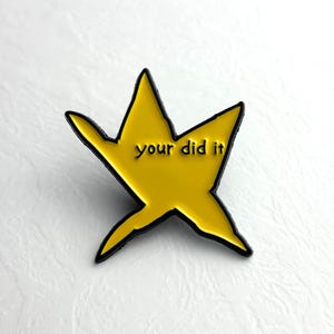 Op de afbeelding: Een gele, stervormige emaille pin met een zwarte omtrek. De pin heeft de tekst "your did it" in zwarte letters in het midden. De pin staat tegen een witte achtergrond.
