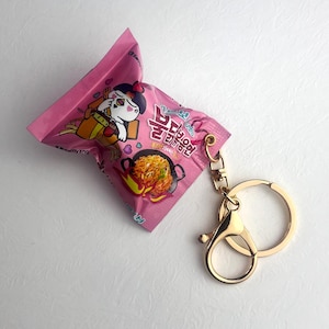Könnte beinhalten: Ein pinkfarbener Schlüsselanhänger in Form einer Packung Instantnudeln. Die Packung hat eine Cartoon-Illustration und Text in einer Fremdsprache. Der Schlüsselanhänger hat einen goldfarbenen Verschluss und Ring.