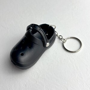 Peut inclure: Un porte-clés en forme de sabot noir miniature. La petite chaussure a une surface texturée, une bride et un porte-clés en métal argenté. Le porte-clés est un article de fantaisie.