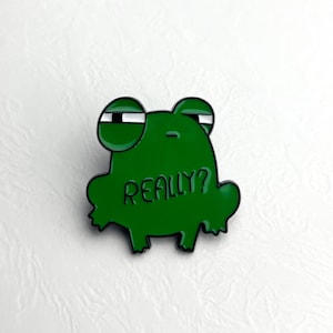 Könnte beinhalten: Ein grüner Emaille-Pin in Form eines Frosches mit dem weißen Wort "REALLY?". Der Frosch hat große Augen und einen fragenden Ausdruck. Der Pin ist vor einem weißen Hintergrund platziert.