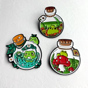 Pode incluir: Três pinos de esmalte com desenhos extravagantes de sapos dentro de garrafas de vidro. Uma garrafa tem um cogumelo vermelho, outra um sapo em um banho e a terceira flores. Cada pino tem uma rolha de cortiça.