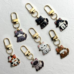 Puede incluir: Una colección de llaveros con forma de gato con cierres dorados. Los llaveros presentan varios diseños de gatos en negro, gris, blanco y marrón, con detalles como rayas y manchas. Cada llavero está unido a un cierre dorado.