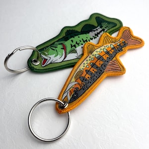 Könnte beinhalten: Zwei bestickte Fisch-Schlüsselanhänger. Einer ist grün mit einem weißen und roten Fischdesign, der andere ist orange mit einem braunen und grauen Fischdesign. Beide Schlüsselanhänger haben silberne Schlüsselringe.