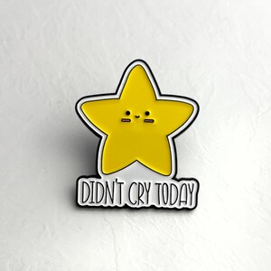 Peut inclure: Une broche en émail en forme d'étoile jaune avec un contour noir et un visage simple composé de deux yeux noirs et d'un petit sourire. Sous l'étoile, une bannière blanche à bordure noire porte le texte noir DIDN'T CRY TODAY.