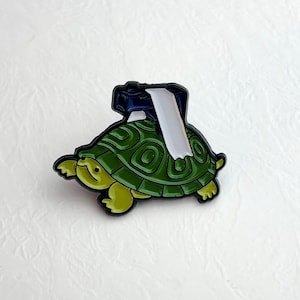 Könnte beinhalten: Ein grüner Schildkröten-Emaille-Pin mit einer dunkelblauen Kamera und weißem Film auf dem Rücken. Die Schildkröte hat einen gelben Kopf und Füße sowie einen gemusterten Panzer. Ein Geschenk für Fotografen und Tierliebhaber.