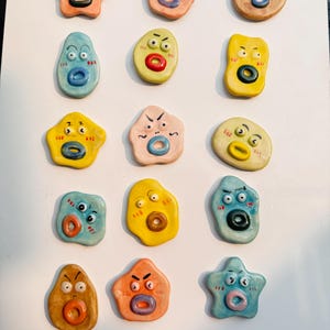 May include: A selection of colourful ceramic magnets, each depicting a cartoon face with a distinctive expression. The magnets feature circular mouths and ring-shaped noses. The shapes vary, including stars and irregular forms.
