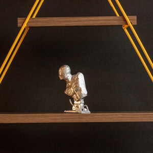Pu&ograve; includere: Una scultura in argento di una testa e spalle umane, con una superficie testurizzata, &egrave; esposta su uno scaffale di legno. Lo scaffale &egrave; sospeso al soffitto da due corde gialle.