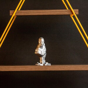 Pu&ograve; includere: Una statuetta argentata di un supereroe con una maschera nera, in piedi su uno scaffale di legno con supporti in corda gialla.
