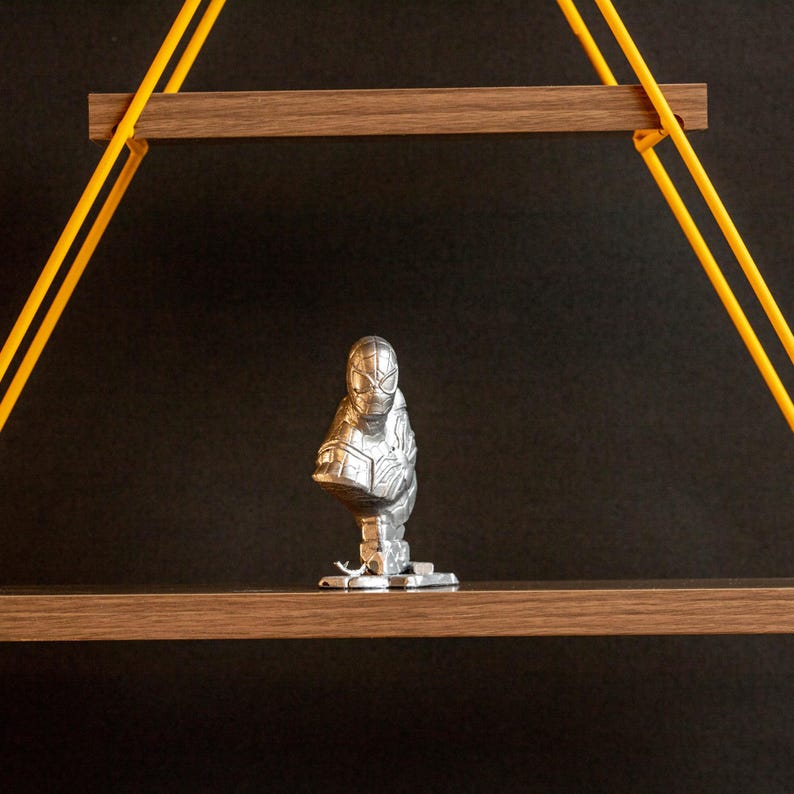 Pu&ograve; includere: Una statuetta argentata di un supereroe con una maschera nera, in piedi su uno scaffale di legno con supporti in corda gialla.