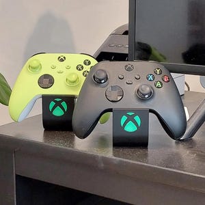 Puede incluir: Dos mandos de Xbox, uno verde lima y otro negro, descansan sobre soportes negros a juego. Los soportes presentan un logotipo verde brillante de Xbox. Los mandos están sobre una superficie oscura, cerca de un monitor.