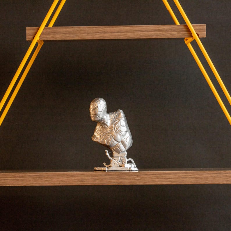 Pu&ograve; includere: Una statuetta argentata di un supereroe, forse Spiderman, in piedi su uno scaffale di legno con corde gialle che pendono dal soffitto.