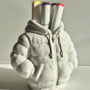 Può includere: Un portapenne in ceramica bianca a forma di giacca invernale imbottita. Quattro pennarelli bianchi con cappucci gialli, blu, viola e rossi sono nel cappuccio. La giacca ha una cerniera e tasche.