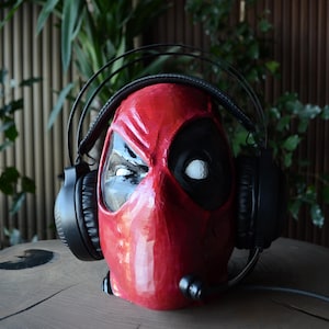 May include: A red Deadpool mask with black eyes and a black microphone attached to the bottom. The mask is wearing black over-ear headphones.