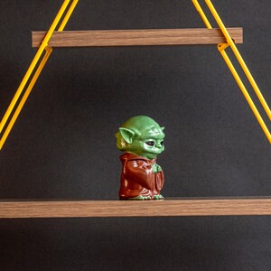 May include: A green and brown figurine of a character from the Star Wars franchise, standing on a wooden shelf with yellow rope supports.
