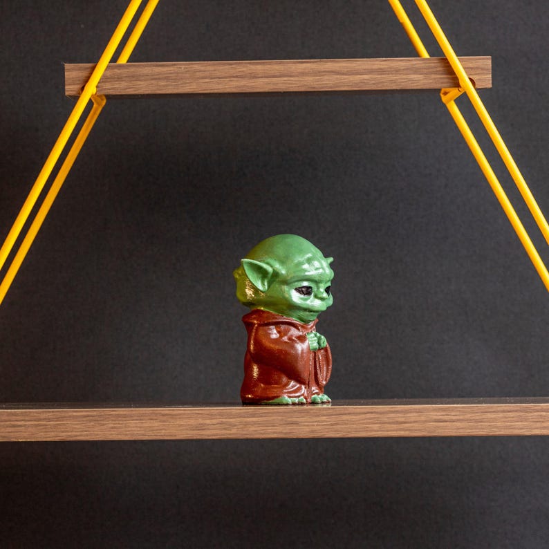 May include: A green and brown figurine of a character from the Star Wars franchise, standing on a wooden shelf with yellow rope supports.