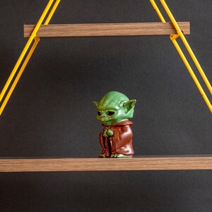May include: A green and brown Yoda figurine standing on a wooden shelf with yellow rope hanging from the ceiling.