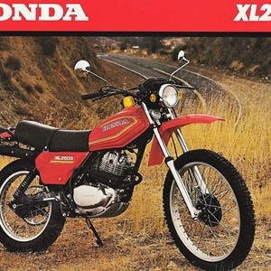 May include: A red Honda XL250S motorcycle is pictured in a field. The motorcycle has a black seat, silver engine components, and a round headlight. The Honda logo is visible on the fuel tank. The background features a winding road and dry grass.