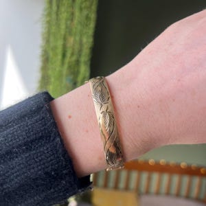 May include: A gold-colored bracelet with an engraved leaf design. The bracelet is a cuff style, approximately 0.75 inches wide, and is worn on a wrist. The background is blurred, with hints of green and other colors.