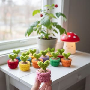 May include: A collection of colorful, crocheted miniature plant pots, each with a heart-shaped green leaf. The pots are in various colors, including red, yellow, orange, and green. A larger potted plant and a mushroom-shaped lamp are in the background.