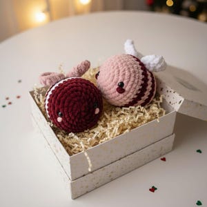May include: Two handmade crochet bee ornaments in a gift box. One bee is burgundy and white, the other is pink and burgundy. The bees have small black eyes and pink accents. The gift box is white with gold stars and filled with shredded paper.