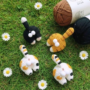 以下が含まれることがあります： 黒、オレンジ、白の猫のお尻の編みぐるみオーナメント。ふわふわの白い尻尾の先と後ろ姿が特徴です。オーナメントは、緑の草の上にデイジーと糸を背景に配置されています。楽しくて風変わりな猫をテーマにしたクラフトです。