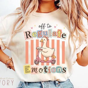 May include: An ivory t-shirt featuring a graphic of a white duck with a pink bow riding a pink scooter. The text reads off to Regulate Emotions in colorful, patterned block letters. The design includes vertical peach stripes and small pink hearts.