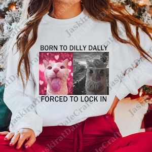 Op de afbeelding: Witte sweatshirt met de tekst "BORN TO DILLY DALLY FORCED TO LOCK IN." De afbeelding toont twee kattenafbeeldingen, één in kleur met harten en de andere in zwart-wit.