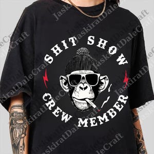 Op de afbeelding: Zwart t-shirt met een aap met een zonnebril en een muts, die een sigaret rookt. De tekst "SHIT SHOW CREW MEMBER" is in een cirkel rond de aap gedrukt, met rode bliksemschichten.