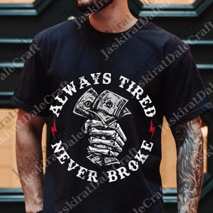 May include: Black t-shirt with a graphic design featuring a skeleton hand holding a wad of cash. The text reads "ALWAYS TIRED NEVER BROKE" in a circular arrangement around the graphic. Red lightning bolt accents are on either side.