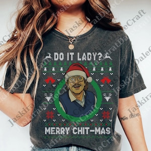 May include: Dark gray t-shirt with a Christmas sweater design. The design includes the text "DO IT LADY?" and "MERRY CHIT-MAS", along with a cartoon image of a man wearing a Santa hat. The shirt has a crew neck.
