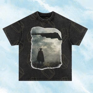 Könnte beinhalten: Dunkelgraues T-Shirt mit einem verwitterten Grafikmotiv einer Figur in einem langen Mantel und Hut, die einen Stab hält, vor einem bewölkten Himmel. Die Grafik hat einen zerrissenen Papiereffekt. Das Shirt hat einen Rundhalsausschnitt und kurze Ärmel.