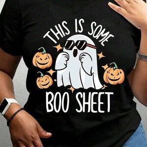 May include: A black short-sleeved t-shirt featuring a white ghost wearing black sunglasses, surrounded by three orange jack-o'-lantern pumpkins and small star shapes. The text "THIS IS SOME BOO SHEET" is printed in white capital letters.