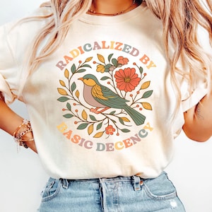 May include: Cream-colored sweatshirt with a colorful graphic. The graphic features a bird perched on a branch surrounded by flowers and leaves. The text "RADICALIZED BY BASIC DECENCY" is arched around the bird.