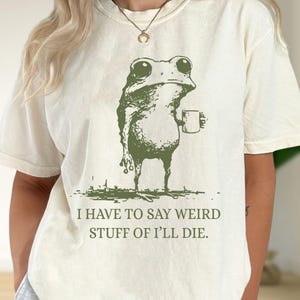 Op de afbeelding: Crèmekleurig T-shirt met een groene illustratie van een kikker die een mok vasthoudt. De kikker heeft grote ogen en een bedachtzame uitdrukking. De tekst hieronder luidt: "I HAVE TO SAY WEIRD STUFF OF I'LL DIE."
