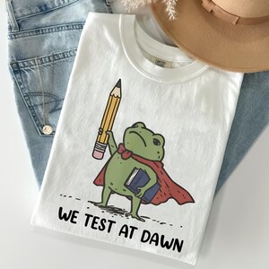 May include: White t-shirt featuring a cartoon frog wearing a cape and holding a pencil and a book. The text "WE TEST AT DAWN" is printed below the frog. The shirt is paired with blue jeans and a tan hat.