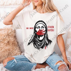May include: Cream-coloured t-shirt with a graphic of a Native American woman with a red hand over her mouth. The text "NO MORE STOLEN SISTERS" is printed in red. The shirt is worn by a person wearing ripped blue jeans.