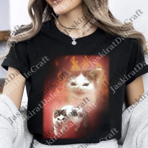 Könnte beinhalten: Schwarzes T-Shirt mit einem Grafikdesign von zwei weißen und braunen Katzen vor einem feurigen Hintergrund. Ein Kätzchen ist von Flammen umgeben. Ein lässiges Kleidungsstück.