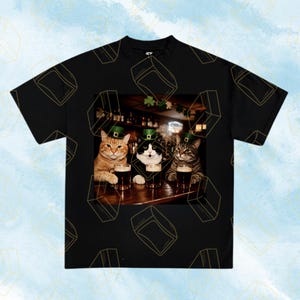 May include: Black t-shirt featuring a graphic of three cats wearing green hats, gathered at a bar with pints of beer. The cats are a ginger tabby, a black and white cat, and a gray tabby. The shirt has a repeating pattern of gold-colored shapes.