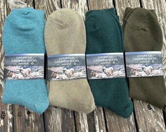 May include: Four pairs of luxurious cashmere socks in shades of teal, tan, dark green, and olive green. Each pair has a label that reads "LUXURIOUS CASHMERE SOCKS MADE IN UK" with an image of sheep. The socks are folded and displayed on a wooden surface.