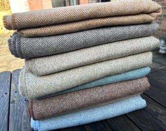 Puede incluir: Una pila de telas plegadas con estampado de espiga en varios colores, incluyendo beige, gris y azul. La tela parece ser un material tejido, posiblemente lana o una mezcla de lana. Los bordes de la tela están cuidadosamente acabados.
