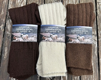 May include: Three pairs of ribbed cashmere socks in shades of brown and cream, each with a label that reads "LUXURIOUS CASHMERE SOCKS. MADE IN UK." The socks are folded and displayed on a weathered wooden surface.