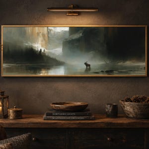 May include: A rectangular landscape painting in a gold frame depicting a misty lake with a moose standing in the water, dark evergreen trees, and towering cliffs. The artwork is illuminated by a horizontal bronze wall light fixture mounted above.