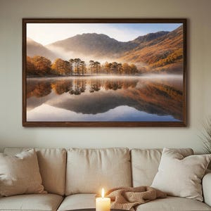 May include: A framed landscape print depicts a serene lake scene with mountains in the background. The water reflects the trees and mountains, creating a mirror image. The artwork is displayed above a beige sofa with pillows, a lit candle, and a blanket.