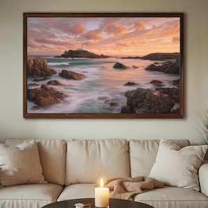 May include: A framed landscape print depicts a coastal scene at sunset. The artwork features a lighthouse on a small island, surrounded by ocean waves and rocky cliffs. The sky is filled with orange and pink clouds, and the frame is a dark brown color.